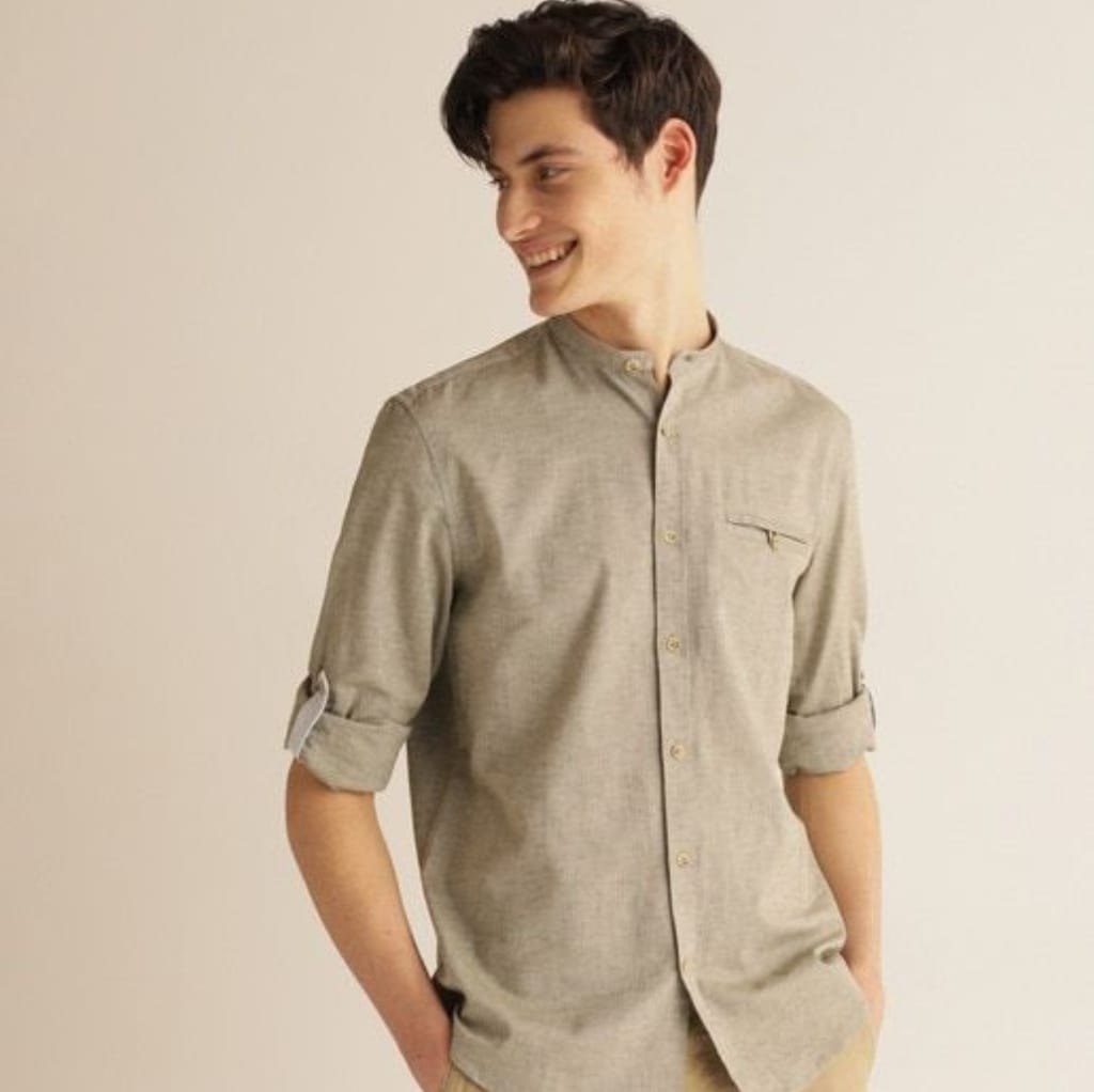 A young man with dark hair smiling and looking to his left, wearing a beige, long-sleeved, button-down shirt with the sleeves rolled up and a stand-up collar.